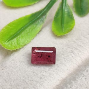 May include: A rectangular, faceted gemstone in a deep pinkish-red hue. The stone features a polished surface with visible dark, natural inclusions scattered throughout its interior, reflecting light with a clear, horizontal highlight across the top.