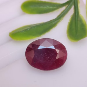 Pode incluir: Uma gema de rubi facetada com corte oval. A pedra vermelha escura está sobre uma superfície branca, com folhas verdes no fundo. As facetas da gema refletem a luz.