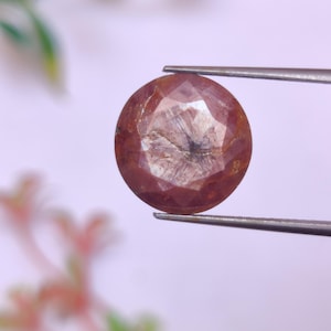May include: A round, faceted, pink gemstone with a darker, almost black, center. The gemstone is held by tweezers against a white background.
