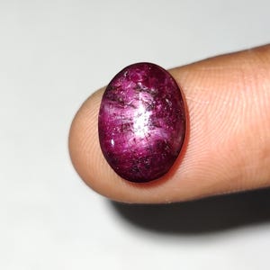 May include: A polished, oval-shaped ruby gemstone. The gem is a deep, saturated magenta color with internal inclusions and a reflective surface. The stone is held between two fingers against a white background, highlighting its color and shape.