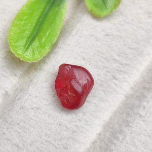 Peut inclure: Une petite pierre précieuse rouge foncé polie, de forme irrégulière et arrondie, avec une surface lisse et légèrement translucide. La pierre présente de subtiles indentations naturelles et des variations de sa couleur cramoisie vive.