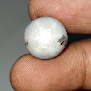 Op de afbeelding: Een klein, bolvormig, zilverkleurig object met zichtbare onvolkomenheden op het oppervlak. Het object wordt tussen twee vingers gehouden, met een neutrale grijze achtergrond. De bol lijkt van metaal te zijn en heeft een licht getextureerd oppervlak.