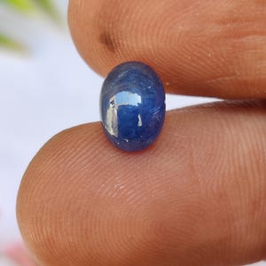 May include: A polished, oval-shaped, deep blue gemstone. The gem has a smooth, reflective surface, with subtle variations in colour. The stone is held between two fingers, showcasing its size and shape. The gem's colour is a rich, saturated blue.