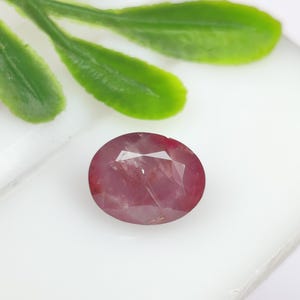 May include: A faceted oval-shaped gemstone with a deep reddish-pink hue. The stone features a polished surface with multiple geometric cuts that reflect light, showcasing internal inclusions and a translucent quality throughout the body.