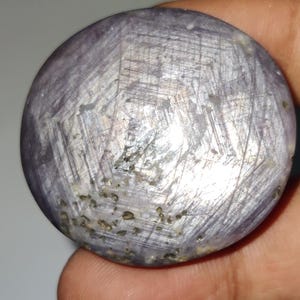 Op de afbeelding: Een ronde, gepolijste edelsteen met een stervormig patroon. De steen heeft een gedempte paarse kleur met een zilverachtige glans. Het oppervlak heeft zichtbare lijnen en kleine donkere insluitsels. De edelsteen wordt in een hand gehouden.
