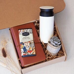 May include: A brown gift box filled with a brown leather journal, a bar of chocolate with the text "Seattle Chocolate Hazelnut Butter Crisp", a white travel mug, and a small potted succulent plant. The box is labeled "BOX 3".