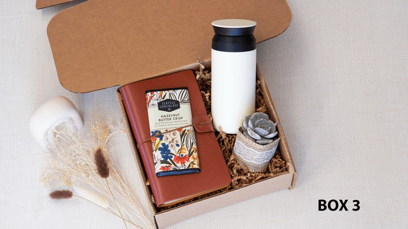May include: A brown gift box filled with a brown leather journal, a bar of chocolate with the text "Seattle Chocolate Hazelnut Butter Crisp", a white travel mug, and a small potted succulent plant. The box is labeled "BOX 3".