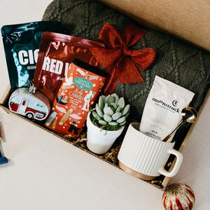 May include: A gift box filled with items for a cozy night in, including a green knitted blanket, a mug, hot chocolate mix, a small potted succulent, and a bar of chocolate with a hot cannoli design.