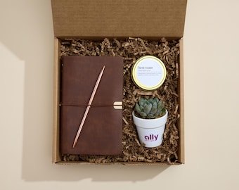 May include: A brown leather journal with a gold pen, a small succulent plant in a white pot with the word "ally" on it, and a small candle with the text "best team" in a gold container. The items are in a brown cardboard box with shredded paper.