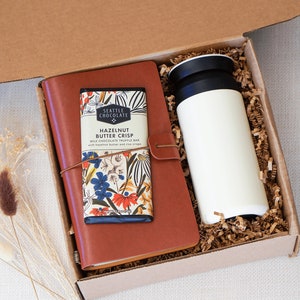 May include: A brown gift box with a brown leather journal, a chocolate bar with floral packaging, and a white travel mug. The box is filled with brown packing paper. The text "BOX 1" is printed on the bottom right corner of the image.