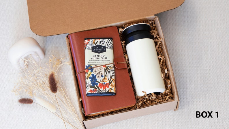 May include: A brown gift box with a brown leather journal, a chocolate bar with floral packaging, and a white travel mug. The box is filled with brown packing paper. The text "BOX 1" is printed on the bottom right corner of the image.