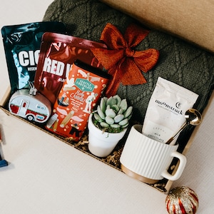 May include: A gift box filled with items for a cozy night in, including a green knitted blanket, a mug, hot chocolate mix, a small potted succulent, and a bar of chocolate with a hot cannoli design.
