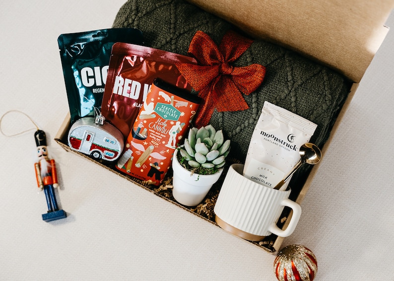 May include: A gift box filled with items for a cozy night in, including a green knitted blanket, a mug, hot chocolate mix, a small potted succulent, and a bar of chocolate with a hot cannoli design.