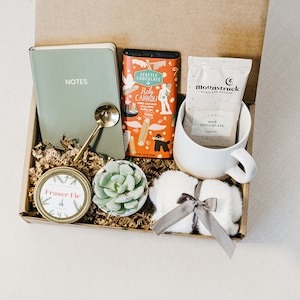 May include: A gift box filled with various items, including a green notebook with the word "NOTES" on the cover, a small potted succulent, a jar of candle, a bar of chocolate, a packet of hot chocolate mix, a white mug, and a white fluffy item with a gray ribbon.