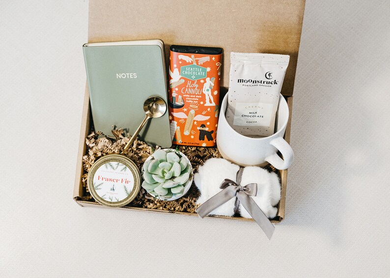 May include: A gift box filled with various items, including a green notebook with the word "NOTES" on the cover, a small potted succulent, a jar of candle, a bar of chocolate, a packet of hot chocolate mix, a white mug, and a white fluffy item with a gray ribbon.