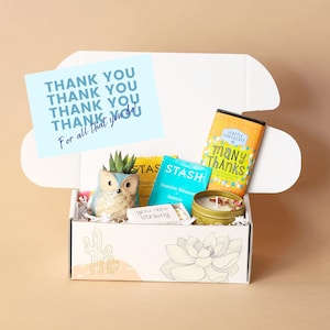 May include: A white gift box with a yellow and blue floral design. The box is open and contains a small succulent plant in a ceramic owl planter, two tea bags, a candle, and a chocolate bar. The chocolate bar has the text "Many Thanks" on it. The box also includes a card with the text "Thank you. Thank you. Thank you. Thank you. For all that you do!"