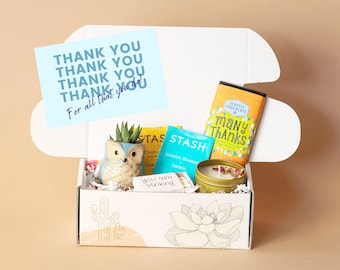 May include: A white gift box with a yellow and blue floral design. The box is open and contains a small succulent plant in a ceramic owl planter, two tea bags, a candle, and a chocolate bar. The chocolate bar has the text "Many Thanks" on it. The box also includes a card with the text "Thank you. Thank you. Thank you. Thank you. For all that you do!"