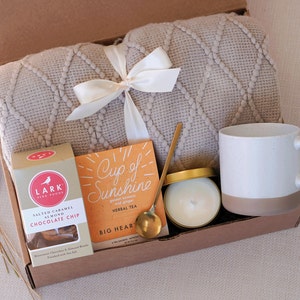 May include: A gift box filled with cozy items, including a beige knitted throw blanket, a white ceramic mug, a small jar of candle, a gold spoon, and a box of herbal tea labeled "Cup of Sunshine".