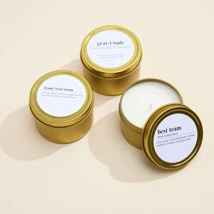 May include: Three gold candle tins with white labels. The labels have the words "grat-i-tude", "best team", and "team that roams" printed on them.