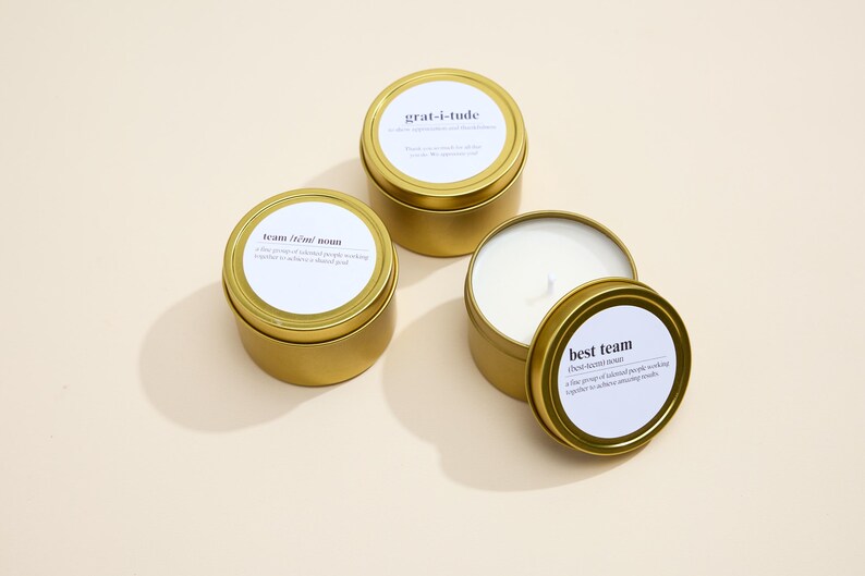 May include: Three gold candle tins with white labels. The labels have the words "grat-i-tude", "best team", and "team that roams" printed on them.
