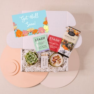 May include: A white gift box filled with a get well soon card, two tea bags, a small succulent plant, a candle, and a bar of chocolate. The card has a blue background with orange and pink flowers. The tea bags are from the brand STASH and the chocolate is from Seattle Chocolate.