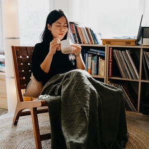 May include: A person sitting in a wooden chair with a green blanket wrapped around them. They are holding a white mug and looking down at it. The chair is in front of a bookshelf filled with books and records.