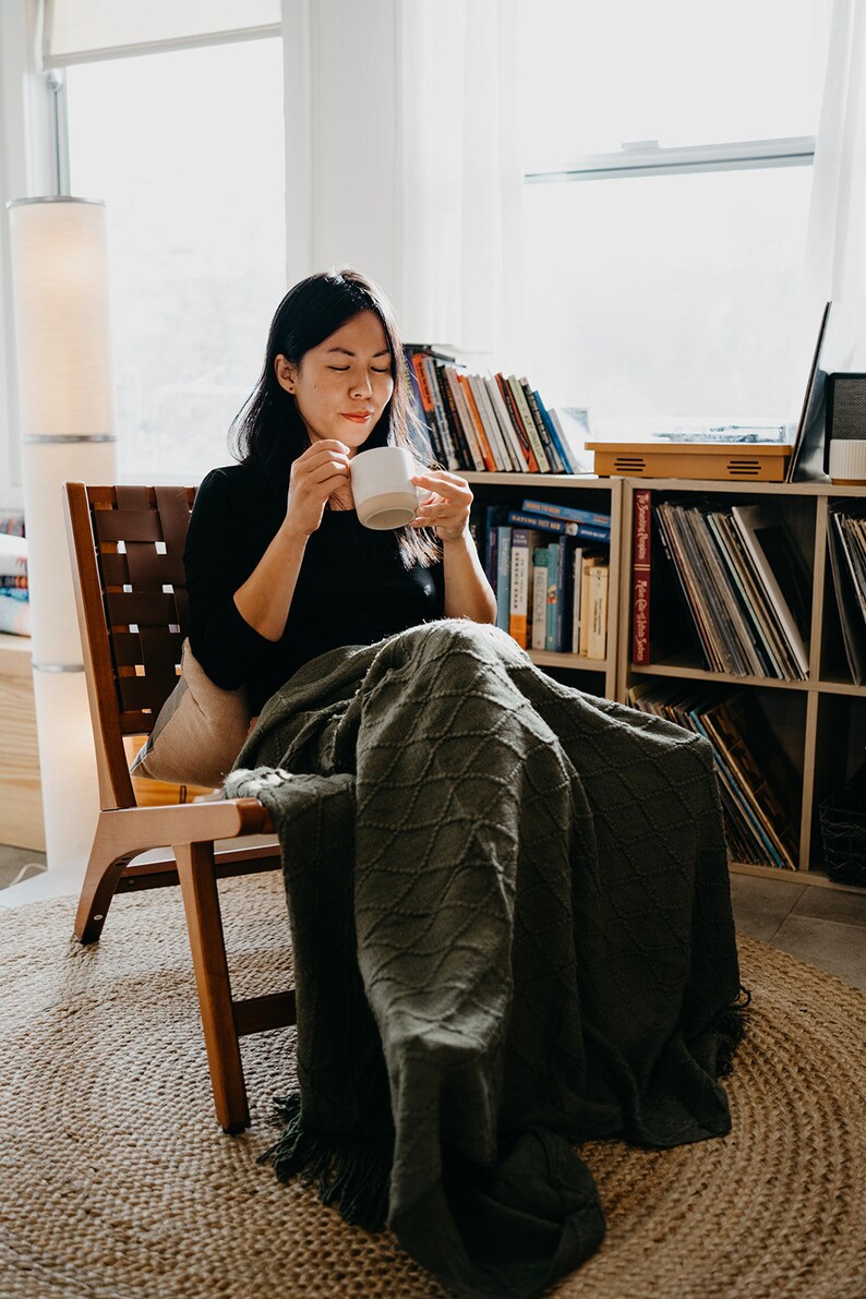 May include: A person sitting in a wooden chair with a green blanket wrapped around them. They are holding a white mug and looking down at it. The chair is in front of a bookshelf filled with books and records.