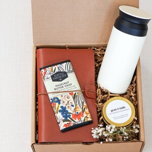 May include: A brown leather journal with a floral design, a bar of chocolate with the text "Seattle Chocolate Hazelnut Butter Crisp", a white travel mug, and a gold jar with the text "gratitude" in a brown cardboard box with shredded paper.
