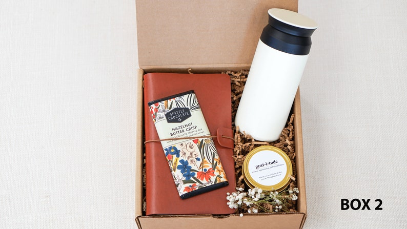 May include: A brown leather journal with a floral design, a bar of chocolate with the text "Seattle Chocolate Hazelnut Butter Crisp", a white travel mug, and a gold jar with the text "gratitude" in a brown cardboard box with shredded paper.