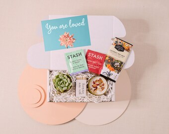 May include: A white gift box with a light pink and white background. The box contains a small succulent plant, a candle, two tea bags, a chocolate bar, and a card that says "You are loved".