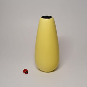 Puede incluir: Un jarrón amarillo liso en forma de lágrima con una abertura estrecha y un borde interior oscuro. Junto al jarrón hay una pequeña figura de mariquita roja y negra. El jarrón tiene un exterior sólido de acabado mate y una base redondeada.