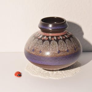 Könnte beinhalten: Eine Keramikvase mit bauchiger Form, mit dunkelbraunem Innenraum und Rand, einem violetten Band und einem gesprenkelten Design in Braun, Weiß und Rosa. Die Vase steht auf einer weißen Spitze, mit einer kleinen roten Marienkäferfigur.