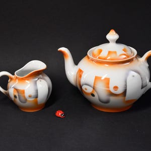 Può includere: Un set da tè bianco con teiera e lattiera con un design astratto arancione e grigio. La teiera ha un coperchio e un beccuccio. La lattiera ha un manico.