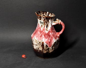 May include: A red and white ceramic pitcher with a brown glaze. The pitcher has a handle and a label that reads "Bay Keramik".