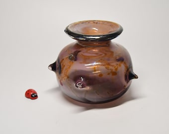 May include: A small, round, iridescent glass vase with a wide, dark-rimmed opening and three protruding glass spikes on its sides. The vase features swirling shades of purple, brown, and gold. A tiny red and black ladybug figurine sits nearby.