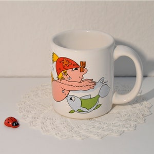 Könnte beinhalten: Weiße Keramiktasse mit einer Cartoon-Illustration einer Person mit roter Mütze, die mit einem Fisch schwimmt. Die Tasse hat einen Henkel.