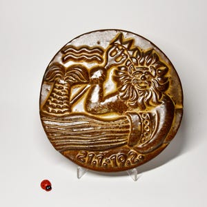 Può includere: Scultura a rilievo rotonda in ceramica marrone raffigurante una figura leonina con corona, onde e una struttura simile a un edificio. La data "27.1.19.2" è incisa. Una piccola coccinella rossa in primo piano.