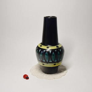 Könnte beinhalten: Eine schwarze Keramikvase mit zylindrischem Hals und bauchigem Körper. Der Körper weist ein Muster aus grünen blattartigen Formen und gelben Akzenten auf. Eine kleine rote Marienkäferfigur befindet sich in der Nähe.