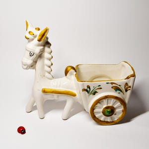 Peut inclure: Un pot de fleurs en céramique blanche en forme d'âne avec une charrette, orné de détails dorés et verts. L'âne a un visage et une crinière détaillés. Une petite figurine de coccinelle rouge est à proximité.