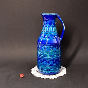 Puede incluir: Una jarra de cerámica azul vibrante con asa, adornada con patrones geométricos en tonos azules y turquesas. La jarra está sobre un tapete de encaje blanco, con una pequeña mariquita roja. La jarra mide aproximadamente 25 cm de alto.