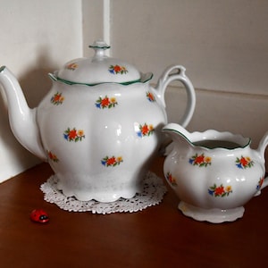 Può includere: Un set da tè bianco con teiera e lattiera con motivi floreali in tonalità arancio, rosso e verde. La teiera ha un bordo verde e un beccuccio sul lato sinistro. La lattiera ha un bordo verde e un manico sul lato destro.