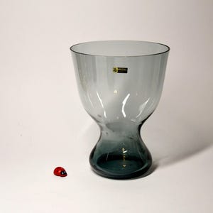 Op de afbeelding: Een heldere glazen vaas met een brede opening en een smalle basis. De vaas is lichtgrijs en heeft een label met de tekst "Kosta Boda Sweden".