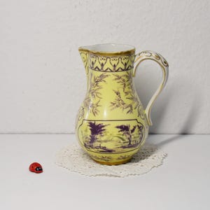 Könnte beinhalten: Gelbe Keramikkrug mit lila Landschaftsdesign und goldenem Rand. Der Krug hat einen gebogenen Griff und steht auf einer weißen Spitze. Ein kleiner roter Marienkäfer befindet sich im Vordergrund.