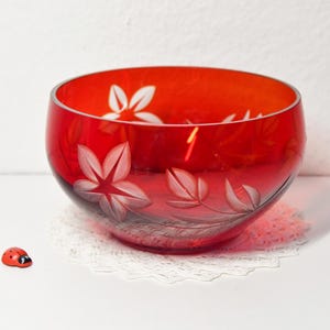 Może przedstawiać: Żywy, czerwony szklany wazon z wytrawionymi wzorami kwiatowymi. Wazon ma białe wzory kwiatów i liści i stoi na białej koronkowej serwetce. Obok wazonu znajduje się mała figurka biedronki.