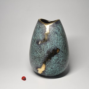 Può includere: Un vaso in ceramica a forma di goccia con una finitura testurizzata maculata color verde acqua e grigio scuro, caratterizzato da una fessura verticale riempita di materiale dorato e un'apertura bordata d'oro, accanto a una piccola coccinella.