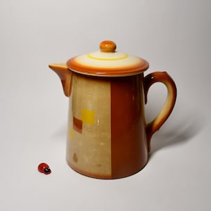 Op de afbeelding: Een vintage keramische koffiepot met een deksel. De pot is voornamelijk bruin met een bruine streep en gele en bruine vierkanten. Het deksel is crèmekleurig met oranje en gele banden. Een klein rood lieveheersbeestje staat ernaast.