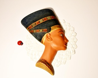 Op de afbeelding: Decoratieve buste van Nefertiti, met een donkerblauwe hoofdtooi en gouden en rode details. De buste heeft een gebruinde teint en staat tegen een witte achtergrond met een kanten kleedje. Er is ook een klein rood lieveheersbeestje aanwezig.