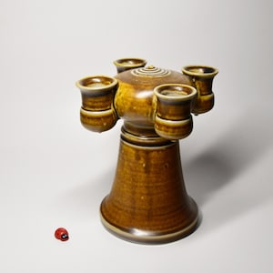 Könnte beinhalten: Ein brauner Keramik-Kerzenhalter mit vier becherförmigen Haltern am oberen Rand. Der Sockel ist ausgestellt, und eine kleine rote Marienkäfer-Figur befindet sich in der Nähe. Der Kerzenhalter hat eine glänzende Oberfläche.