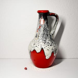 Może przedstawiać: Ceramiczny wazon z czerwoną podstawą, nakrapianym czarno-białym korpusem oraz czerwonym rantem i uchwytem. Wazon ma dołączoną metkę. Wazon stoi na białej powierzchni.