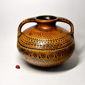 Puede incluir: Un jarrón de cerámica marrón con dos asas y una superficie texturizada. El jarrón está decorado con patrones geométricos negros. Una pequeña mariquita roja está al lado del jarrón.